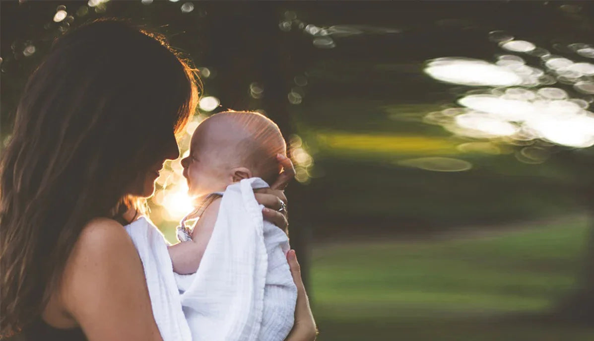 Mother holding baby wrapped in white cotton blanket outdoors, sunlight in background, allergy-free apparel