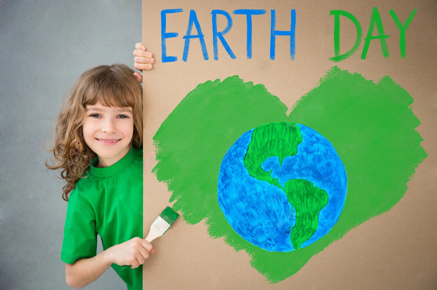 Smiling child in green shirt holding Earth Day sign with painted globe and green heart.