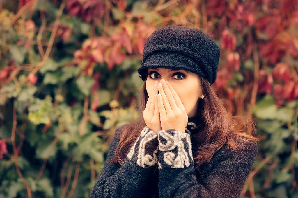 Woman in allergy-free fall clothing outdoors, covering face with hands, surrounded by autumn leaves