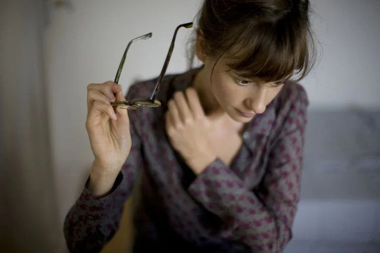Woman in patterned cotton top holding glasses, touching neck, in softly lit indoor setting.