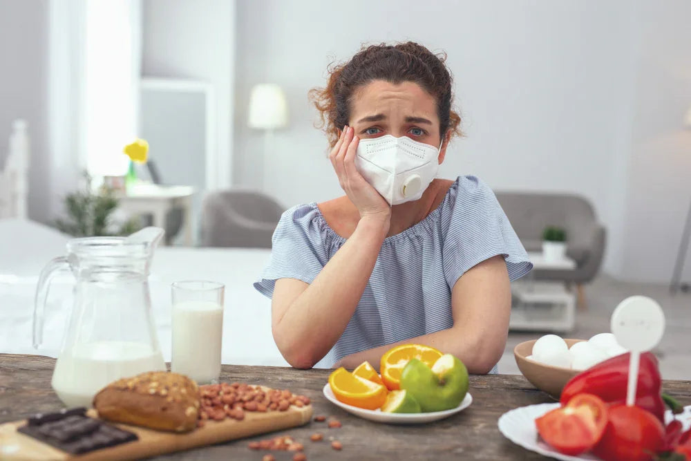 Woman wearing a protective mask, sitting at a table with common food allergens, looking concerned.