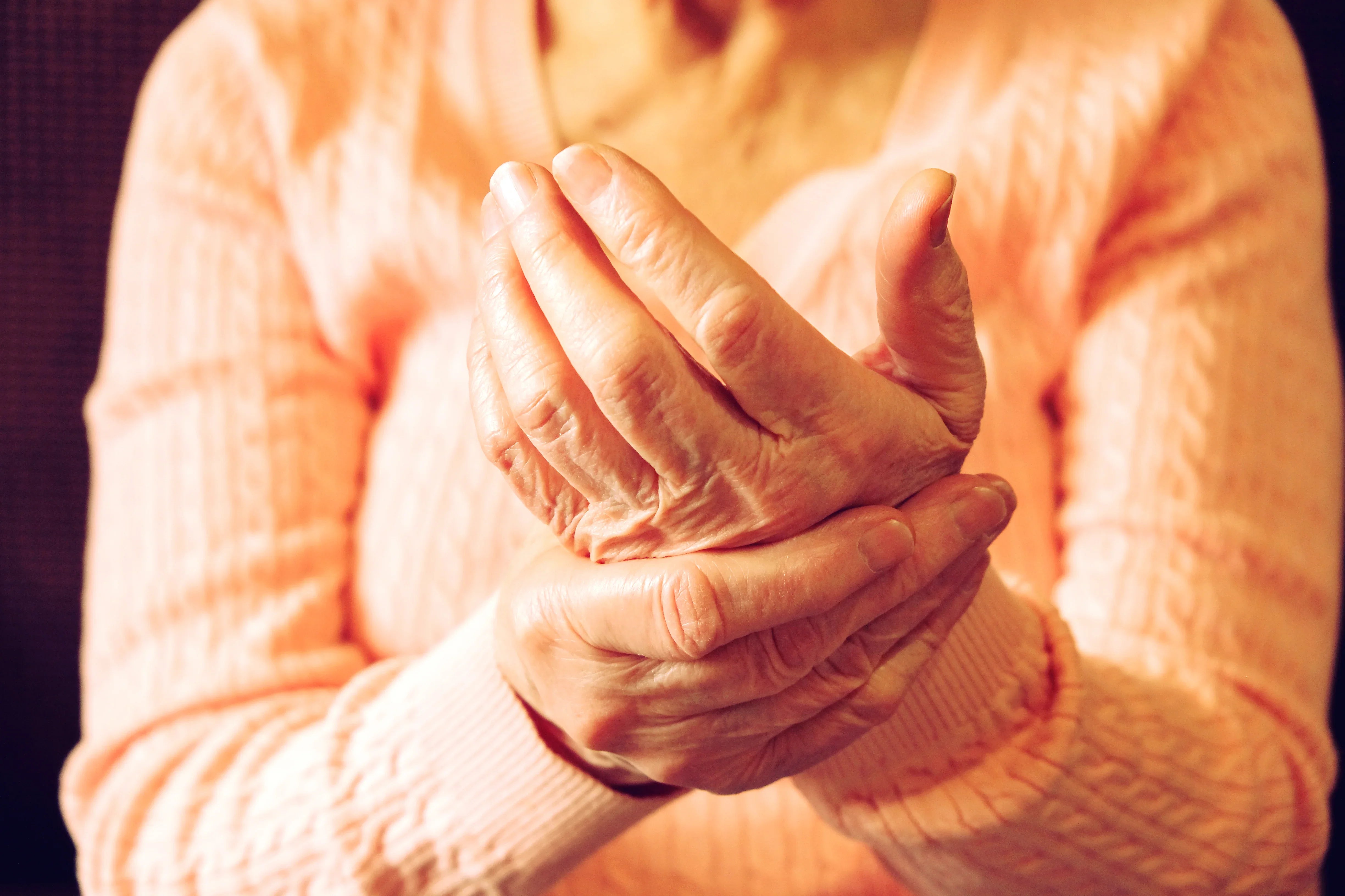 Elderly woman in soft peach sweater gently massaging her hand, sensitive skin care concept