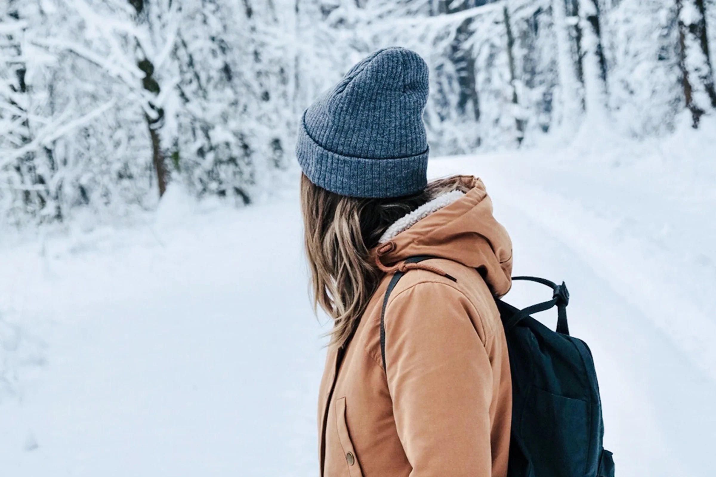 Person in tan winter jacket and gray beanie walking in snowy forest