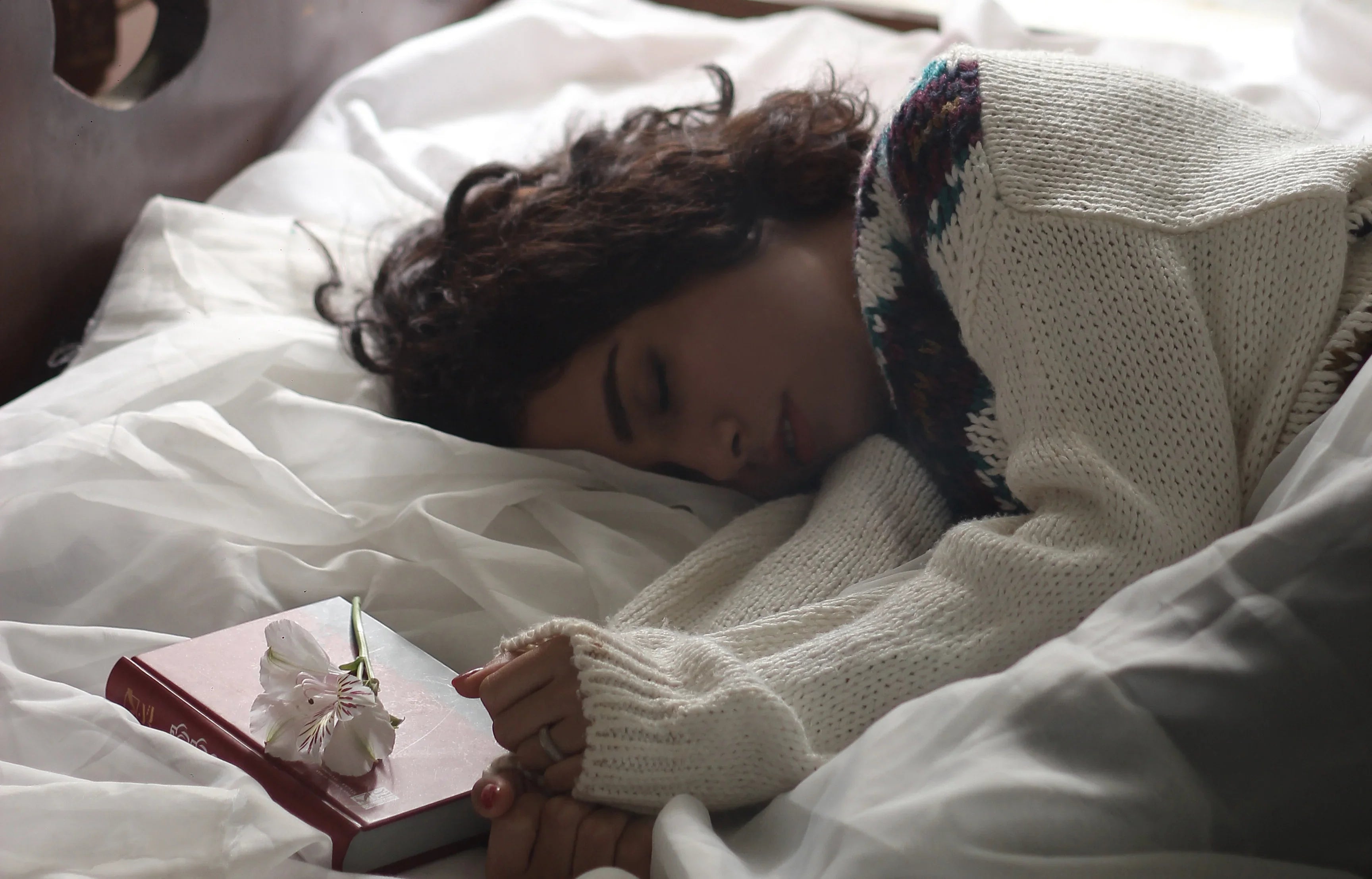 Woman in a cozy knit sweater resting on a bed with a book and flower, soft natural light