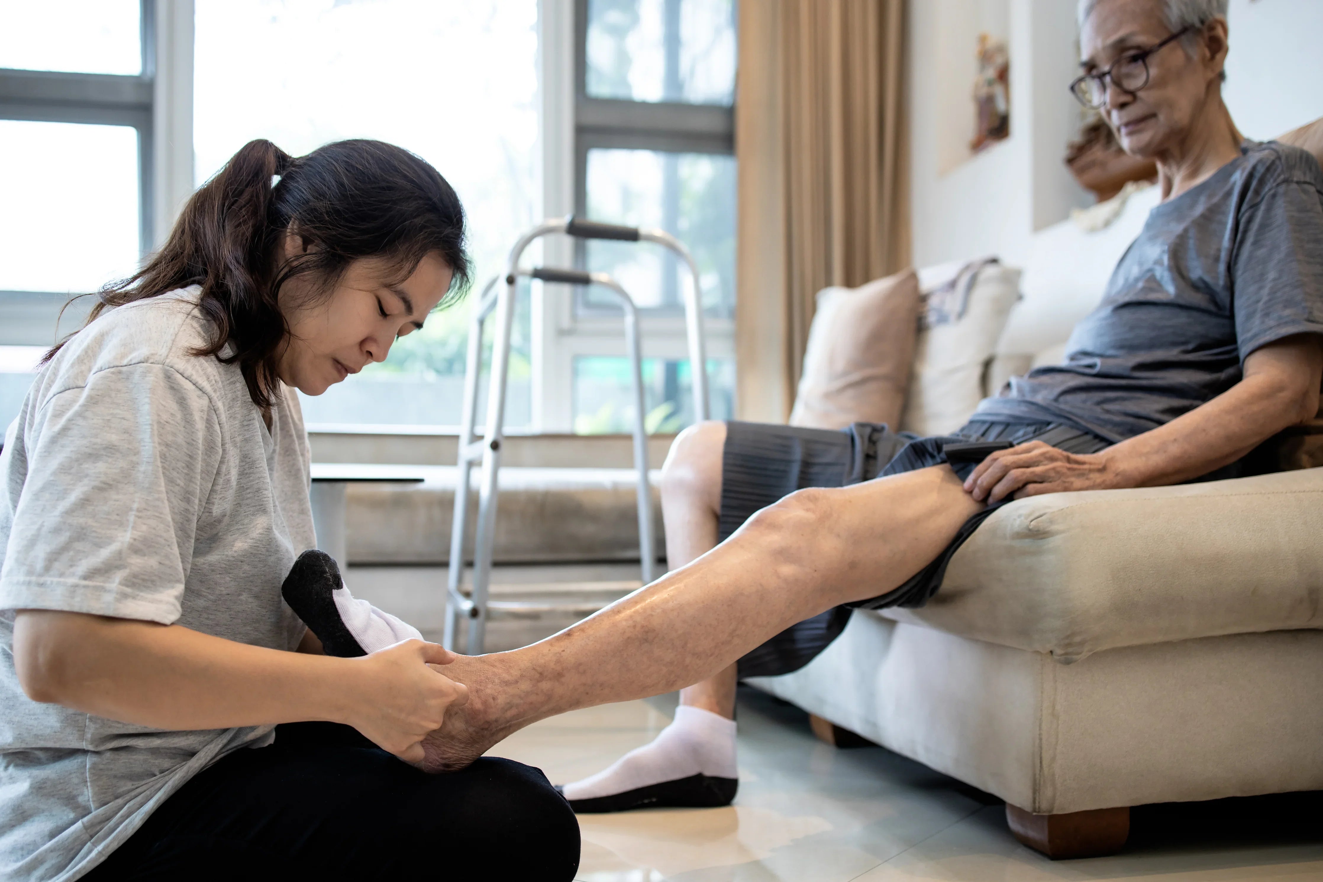 Daughter helps elderly man put on hypoallergenic cotton socks in a bright living room.