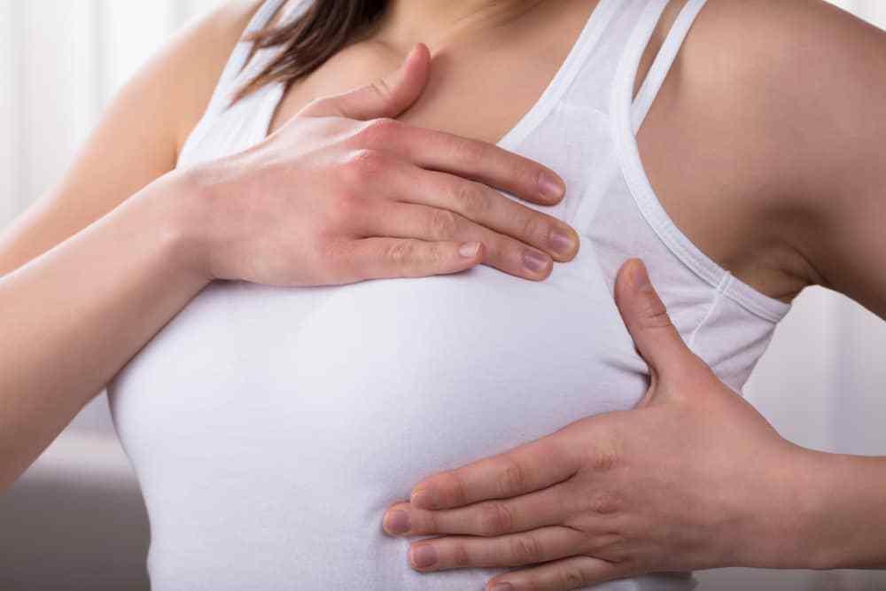 Woman in white cotton tank top performing self-breast massage, hands on chest.