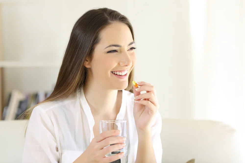 Smiling woman in white shirt holding supplement capsule and glass of water indoors