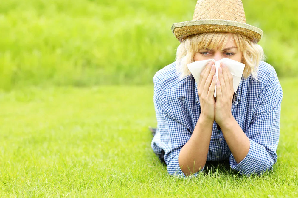 Woman in blue checkered shirt sneezing into tissue on green grass, outdoor allergy concept