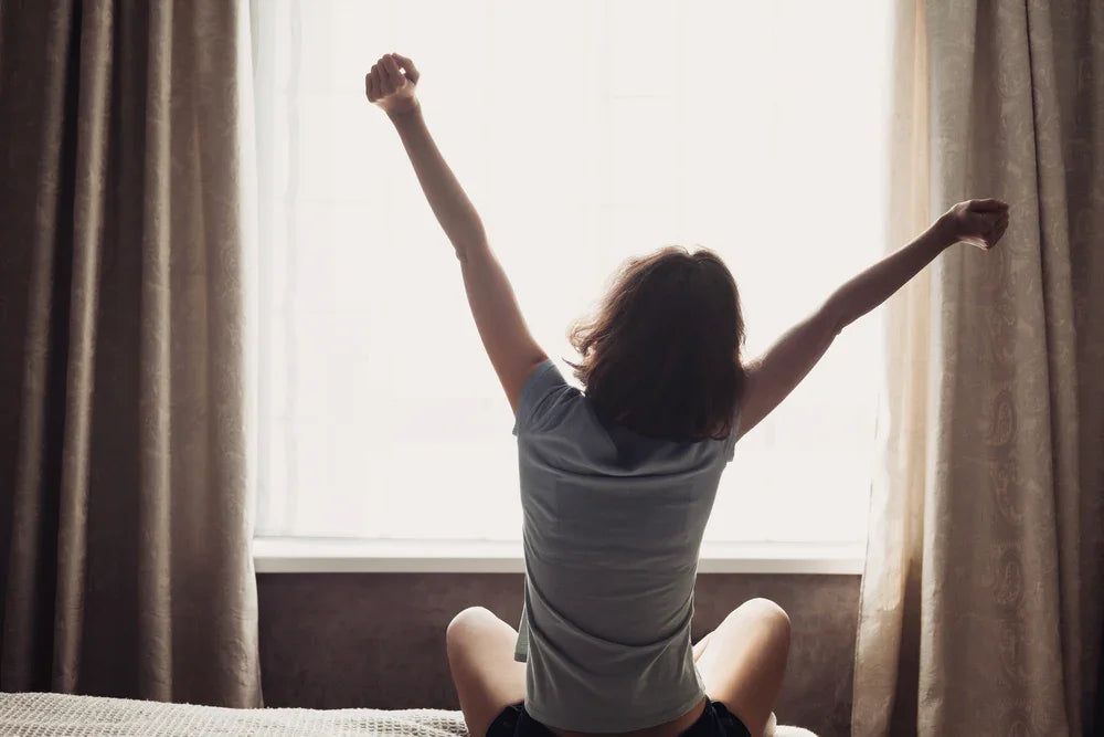 Woman in allergy-free cotton shirt stretching on bed by window, natural light, Cottonique