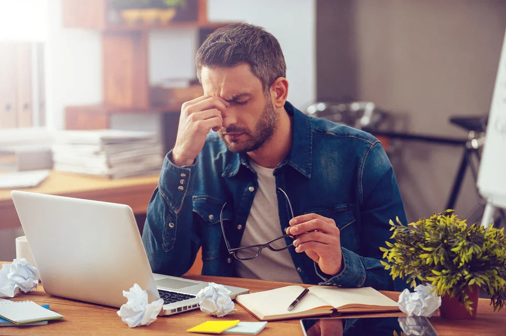 Stressed man in denim shirt at desk with laptop, notebook, glasses, and crumpled paper