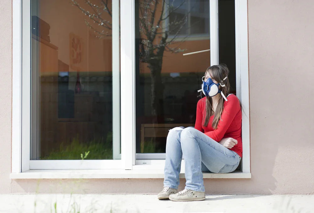 Woman with allergy mask and casual cotton clothing sitting by window, sensitive skin protection