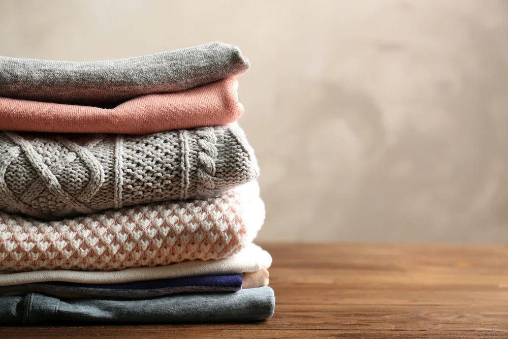 Stack of folded organic cotton sweaters and shirts on wooden table, soft neutral colors