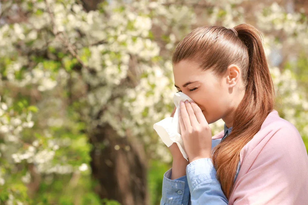 Woman outdoors with tissue sneezing, allergy symptoms, surrounded by blooming trees