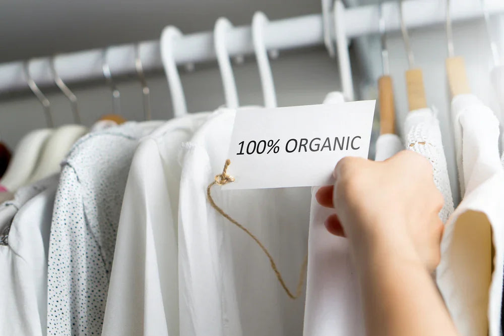 Hand holding 100% organic tag on white cotton shirts hanging in closet, allergy-friendly clothing