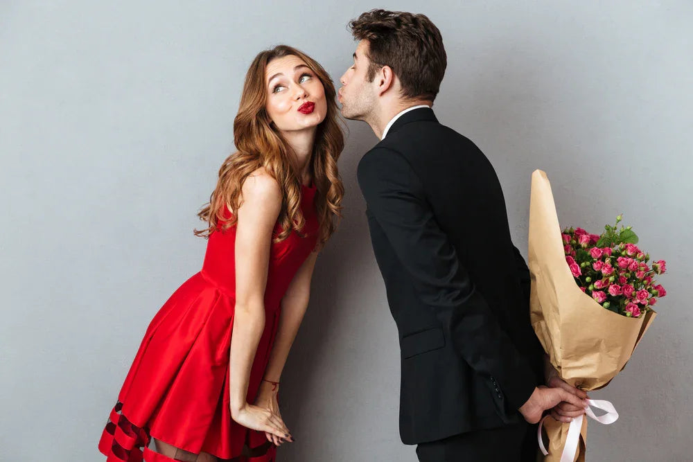 Man in suit holding bouquet behind back leans to kiss woman in red dress, gray background