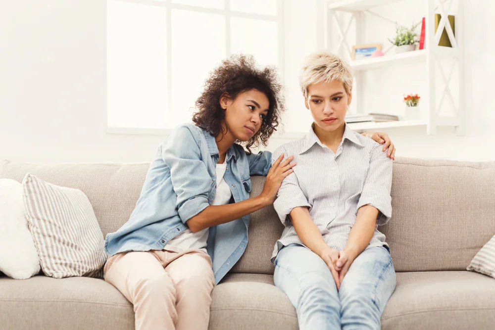 Two women sitting on a couch, one comforting the other in a bright living room