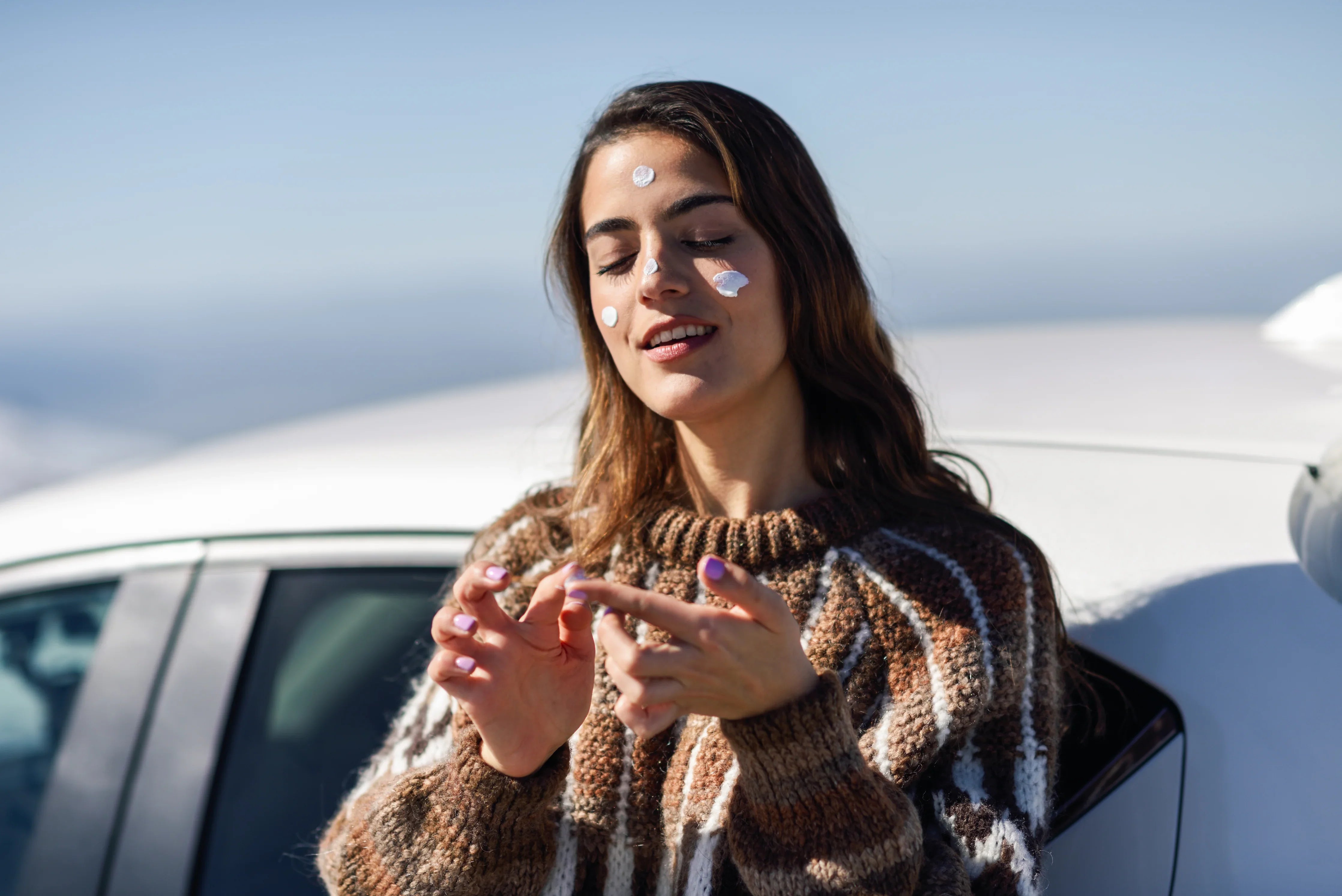 Woman in a hypoallergenic sweater applies cream to face outdoors near a car