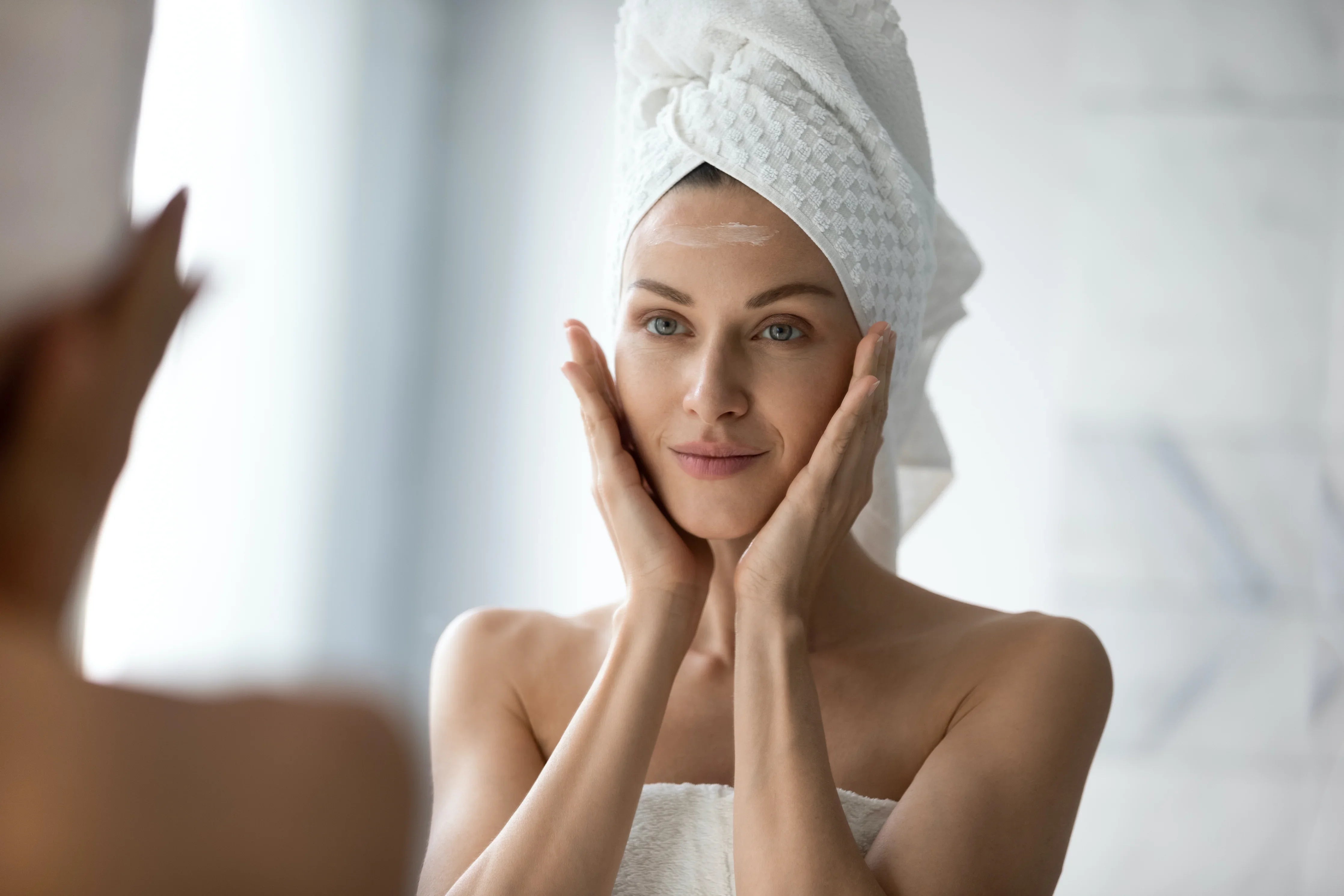 Woman with towel wrapped on head, touching face, wearing soft cotton towel, in bathroom mirror