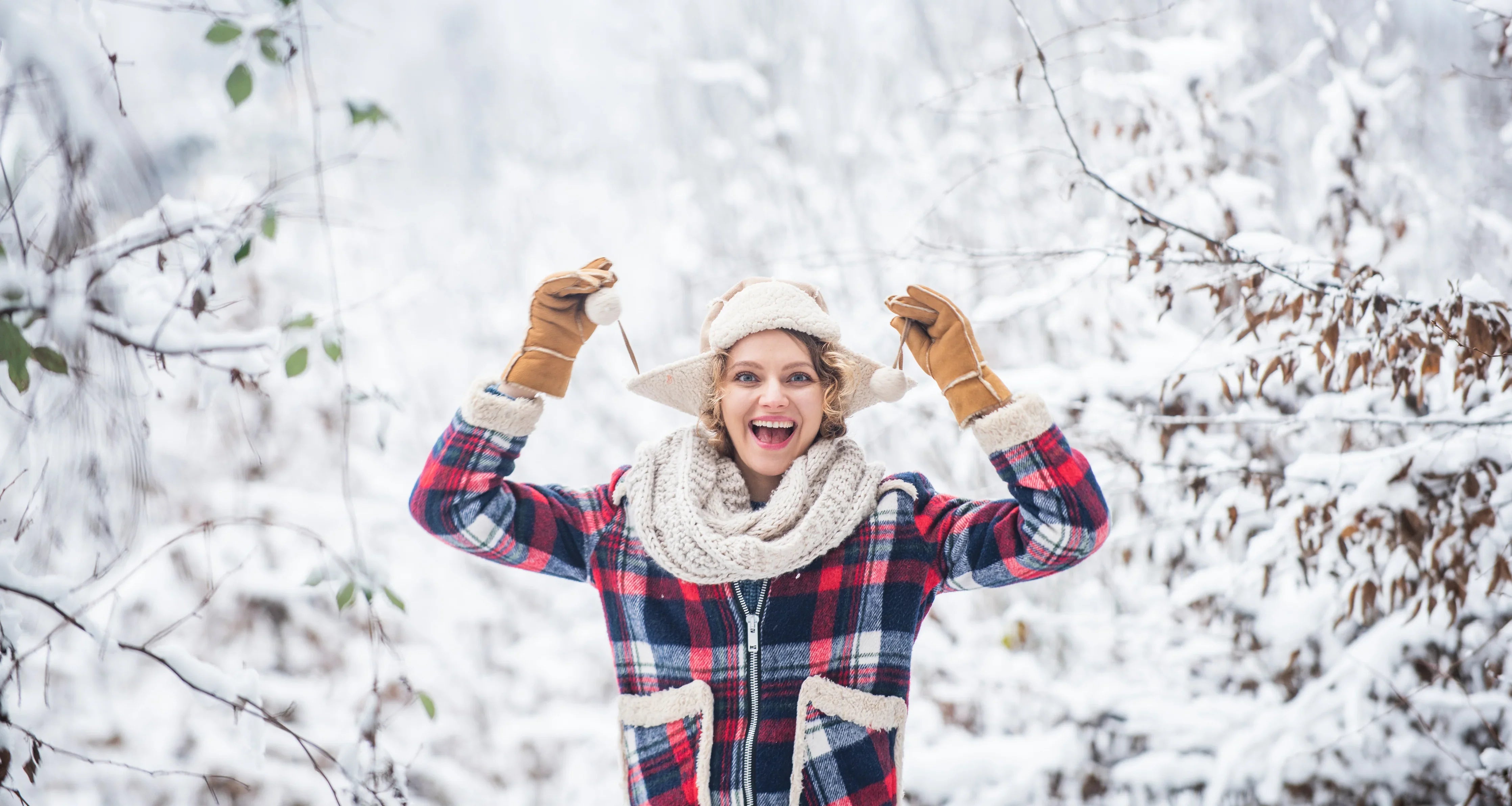 Woman in plaid winter coat, knit scarf, gloves, and hat enjoying snowy forest outdoors