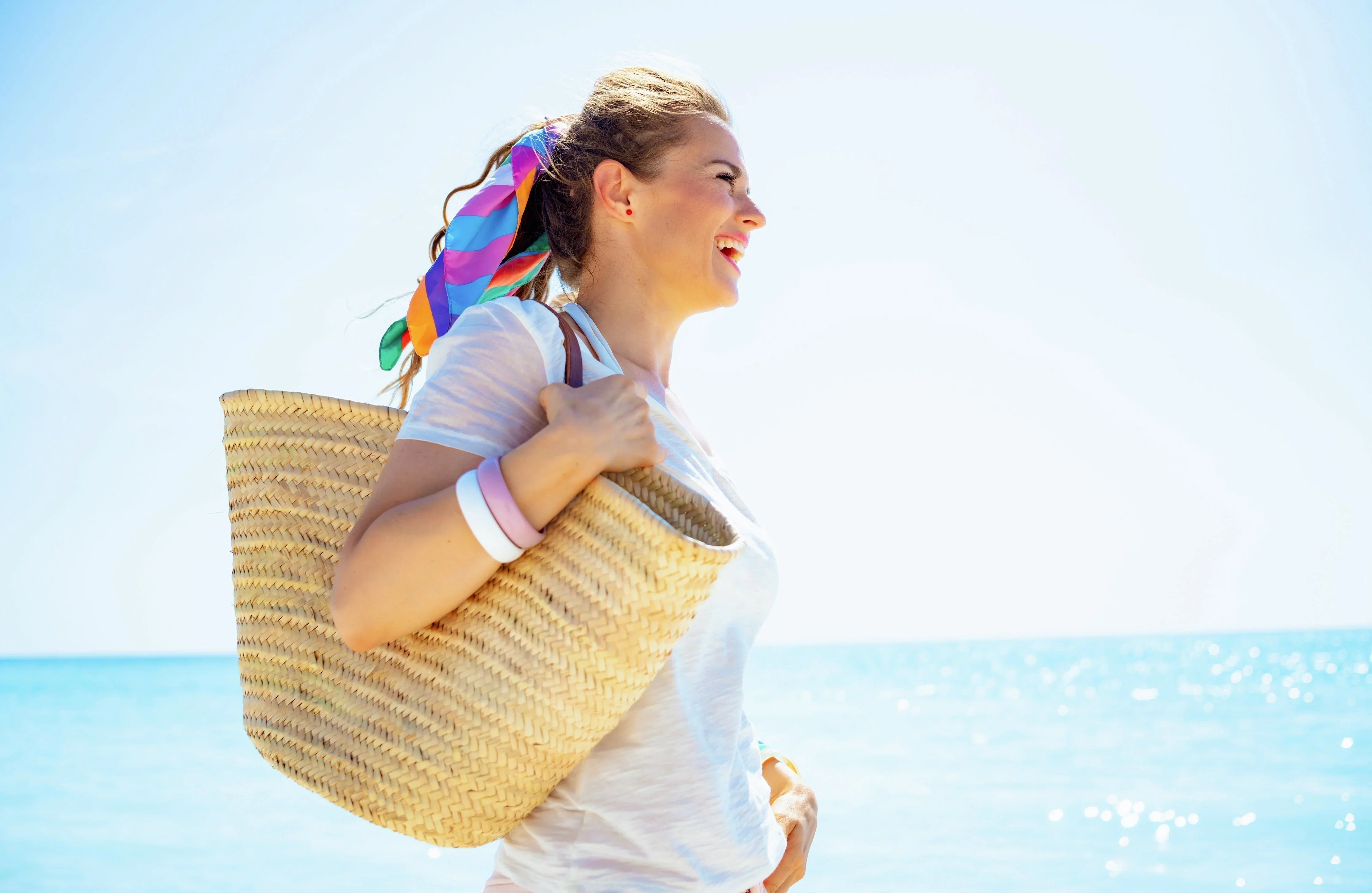 Smiling woman in white t-shirt with woven beach bag and colorful headscarf by the sea