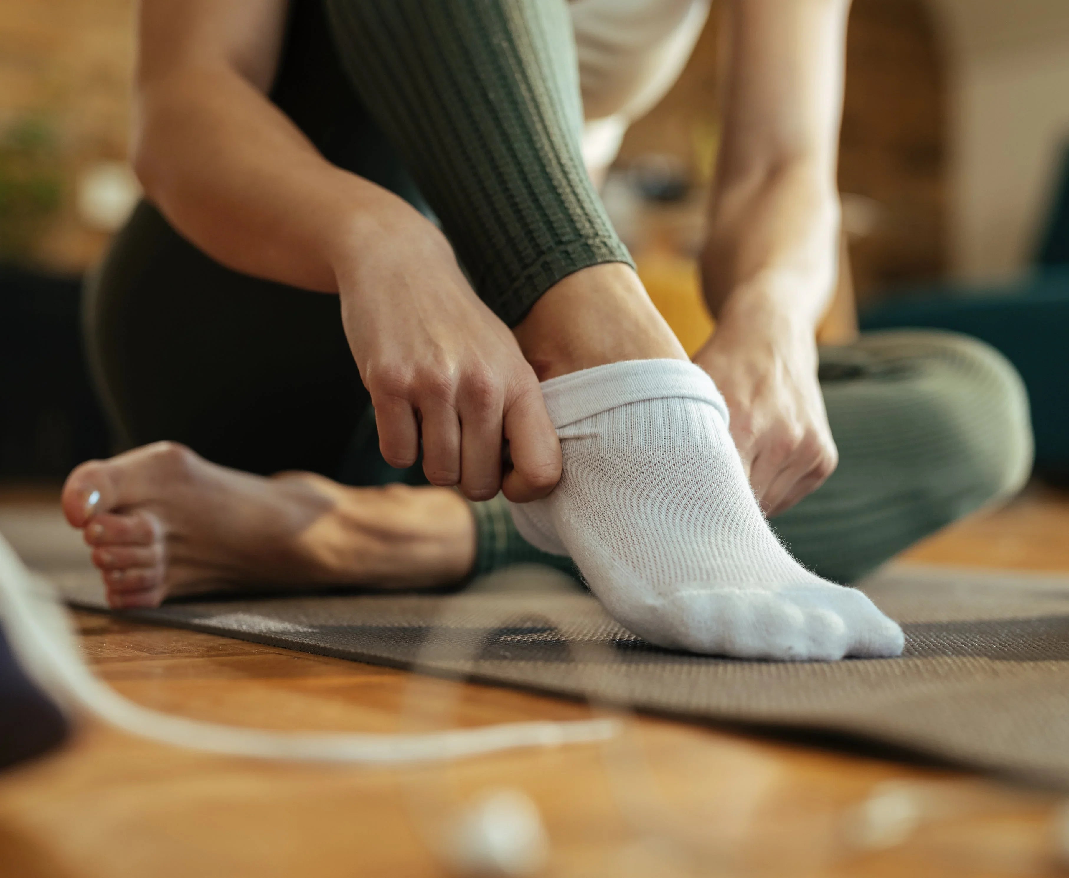 Person putting on white hypoallergenic cotton socks, sitting on a yoga mat indoors