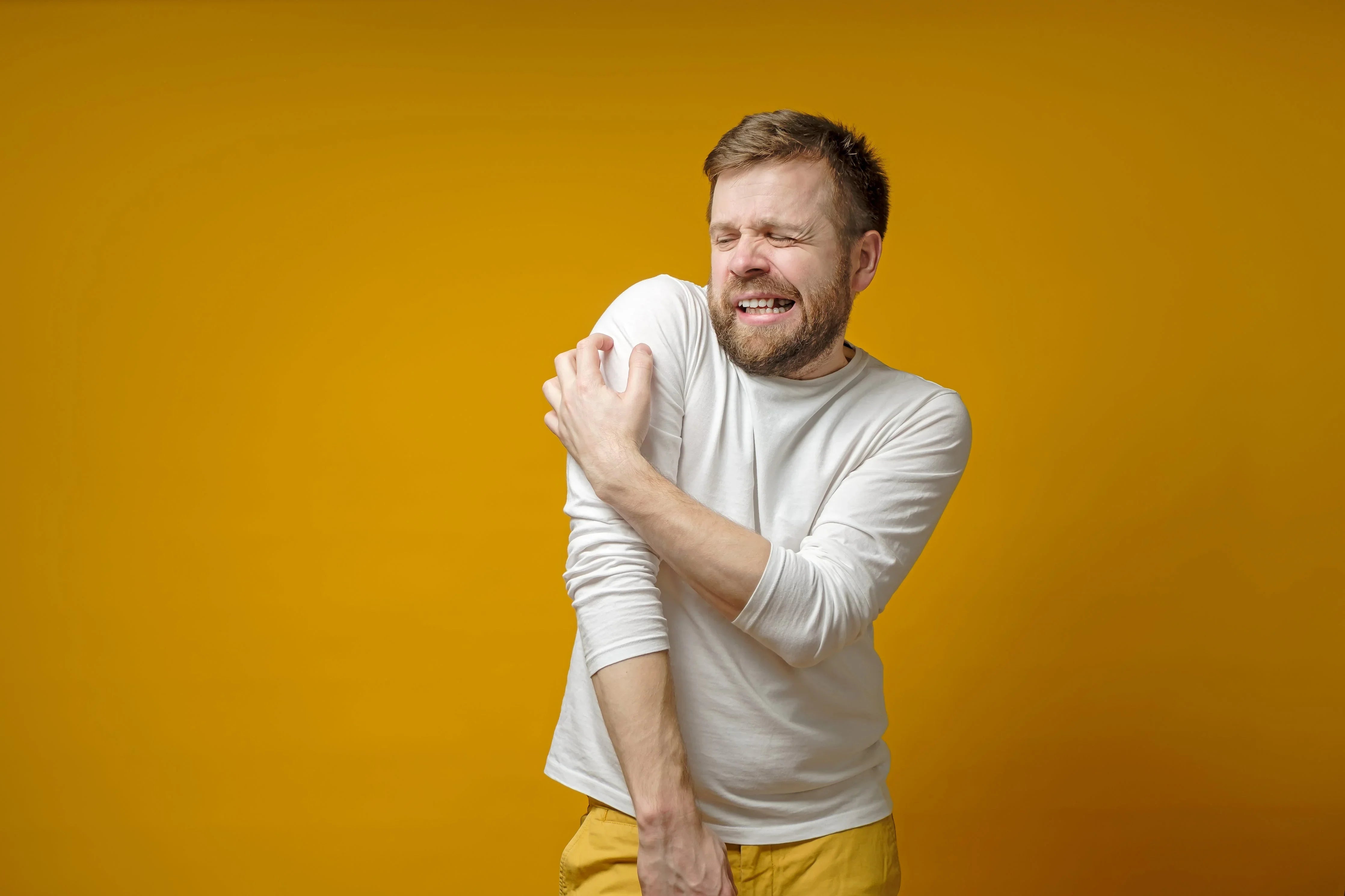 Man in white long sleeve scratching arm, showing skin irritation, on yellow background