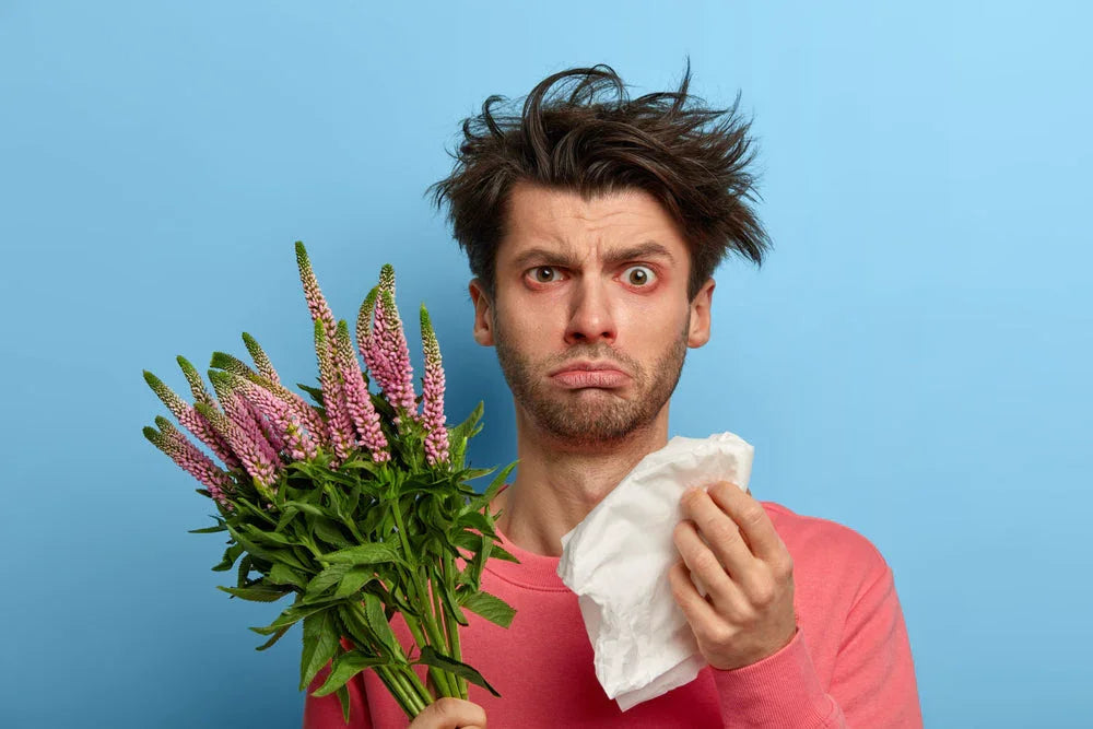 Man with red eyes holding flowers and tissue, showing allergy symptoms, blue background