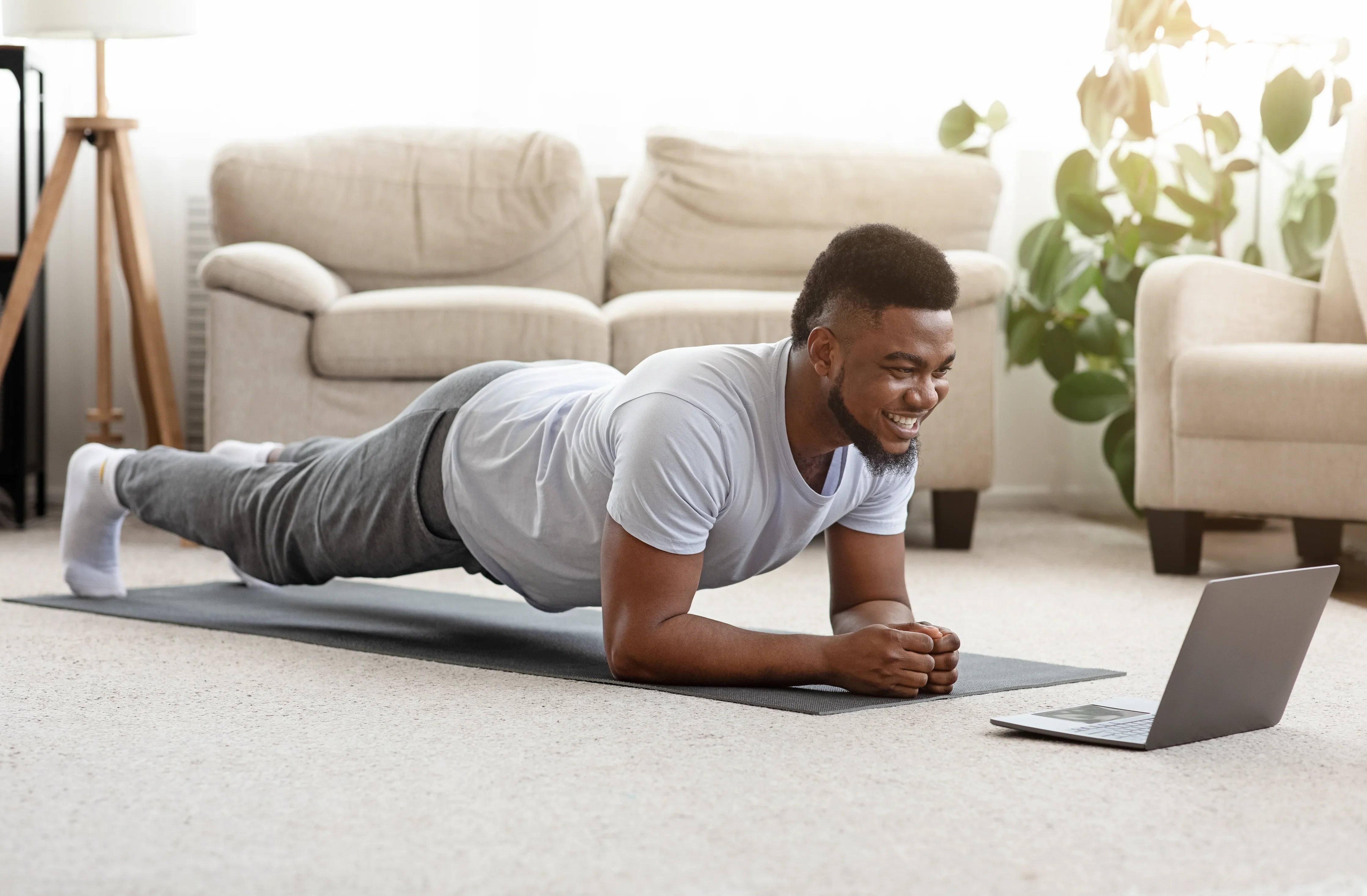 Man in organic cotton t-shirt and sweatpants doing a plank on yoga mat at home with laptop