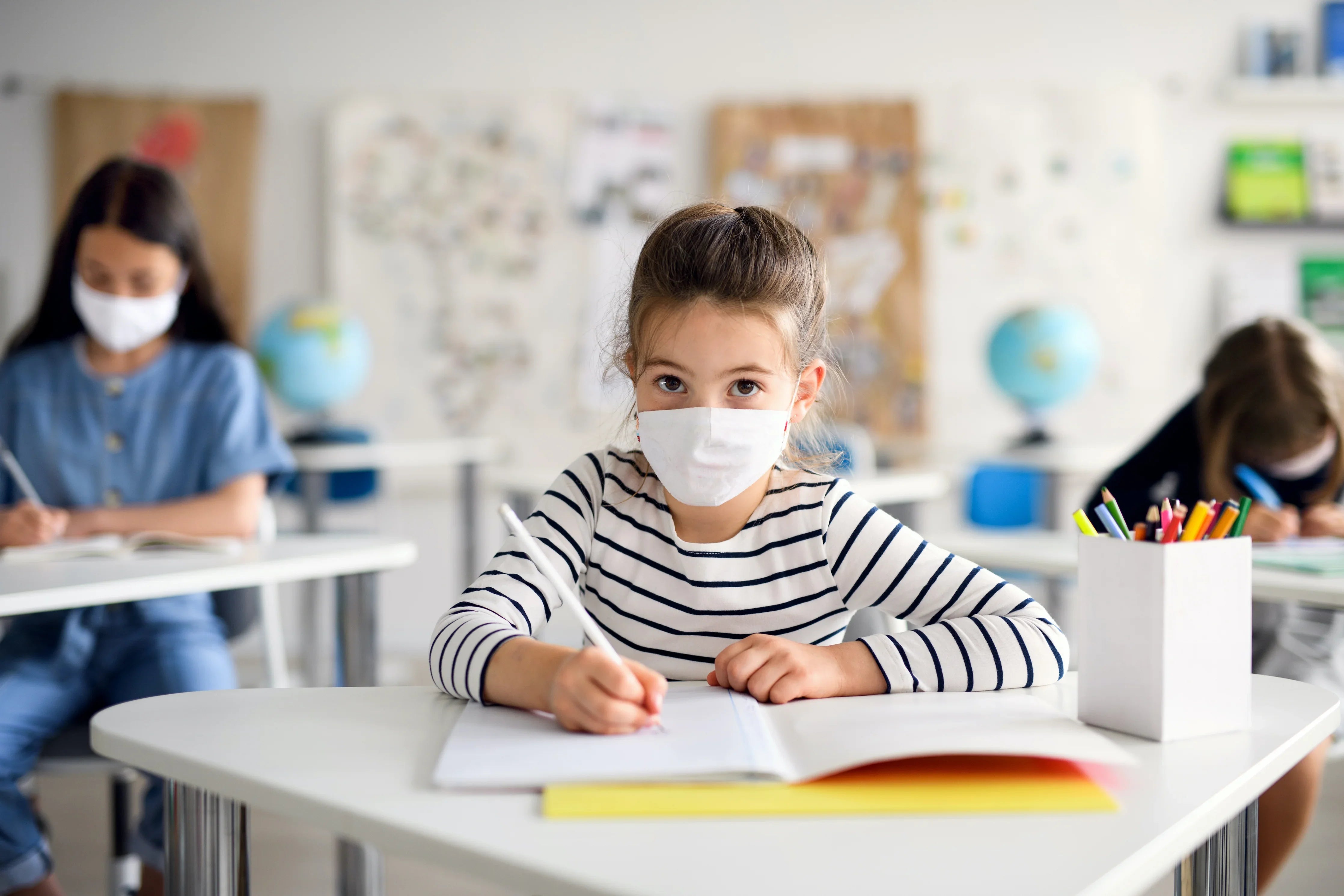 Young girl in striped shirt wearing a cotton face mask, writing at a classroom desk.
