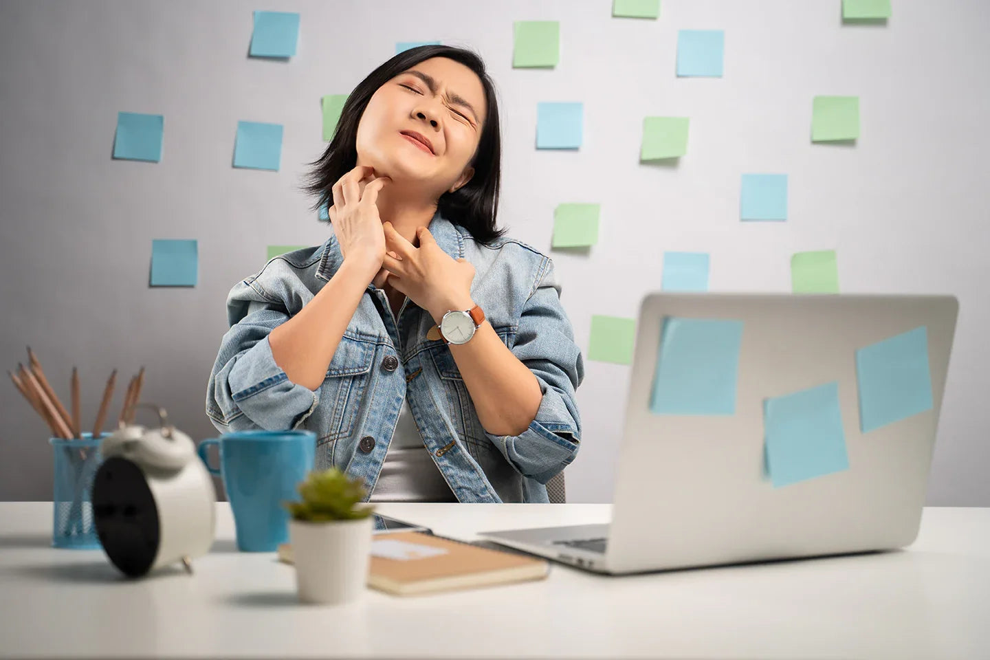 Woman in denim jacket scratching neck, showing skin irritation, sitting at desk with laptop and sticky notes, representing sensitive skin or eczema discomfort.