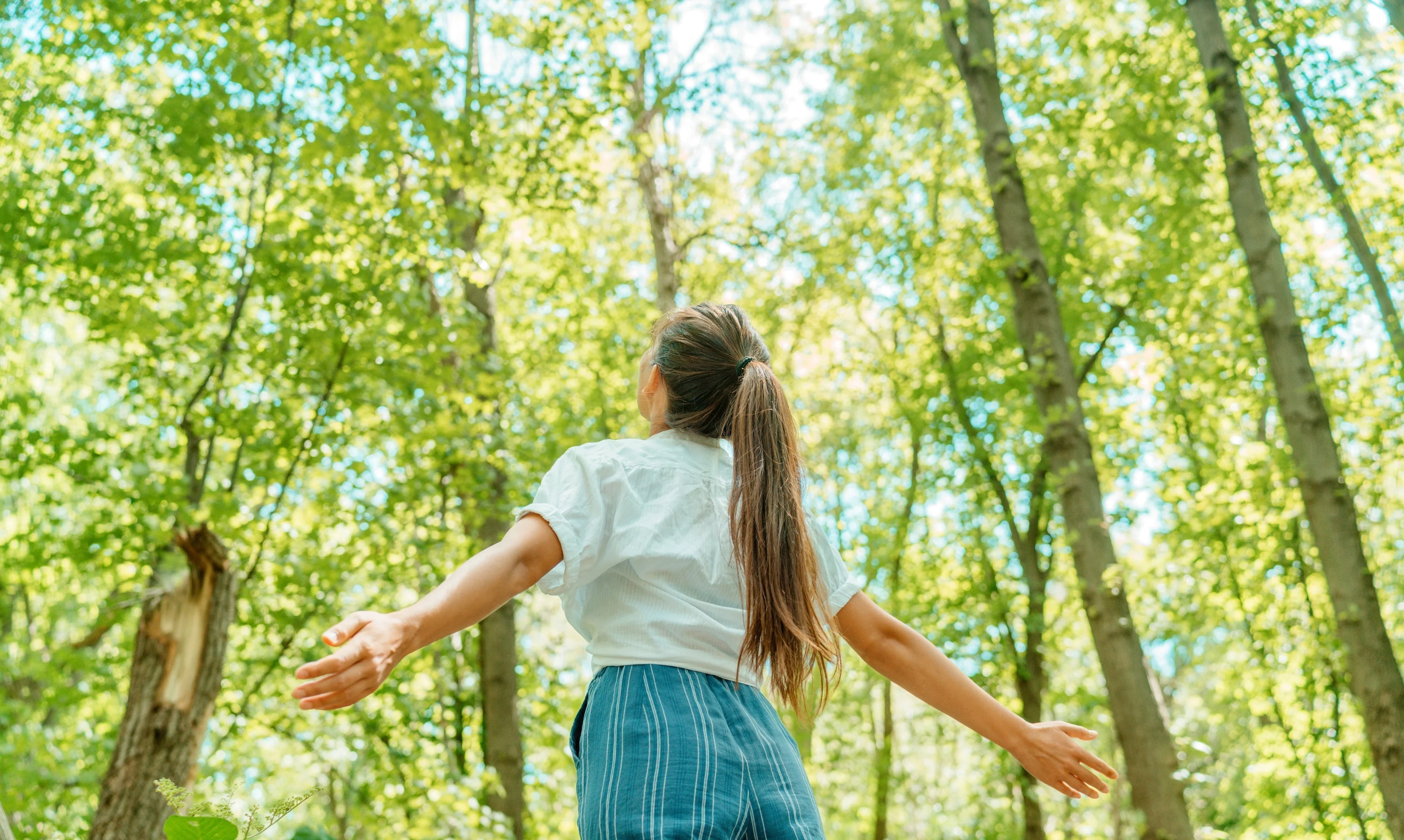 Woman in white cotton shirt and striped pants standing in sunlit forest, arms outstretched