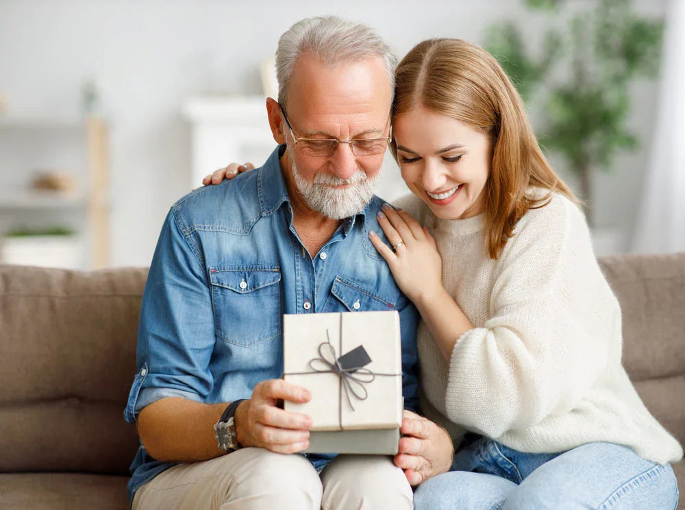 Smiling woman gives older man a wrapped gift box on a cozy sofa, both wearing casual cotton clothing.