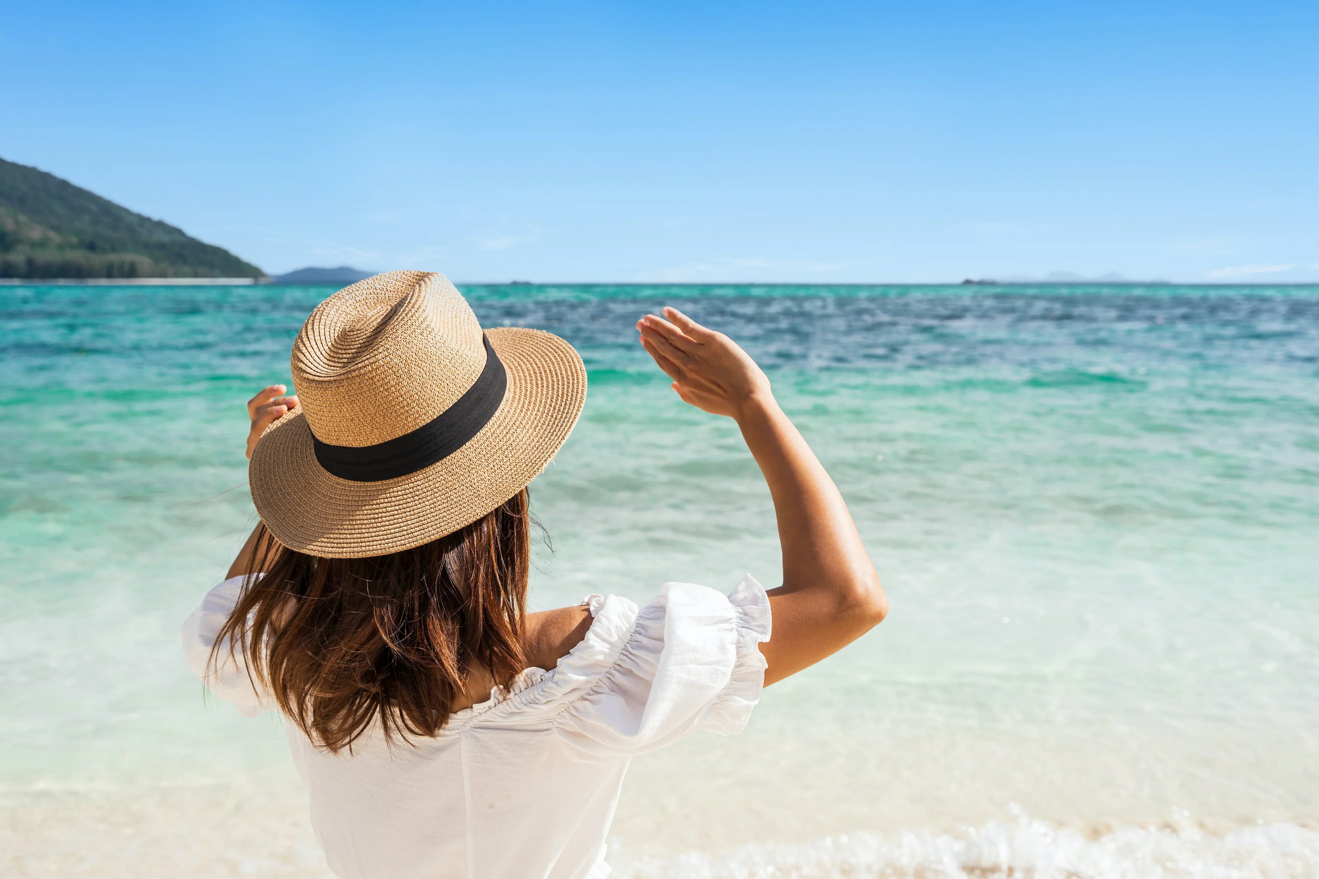 Woman in white cotton blouse and sun hat standing on beach by clear blue sea