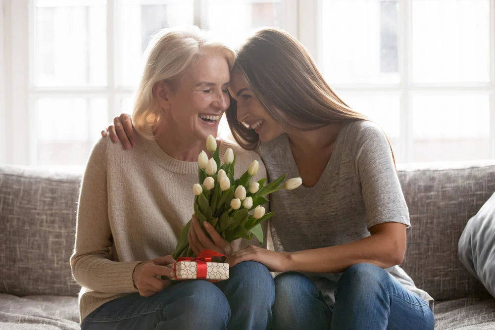 Happy older woman and young woman on couch with white tulips and a gift box, smiling.
