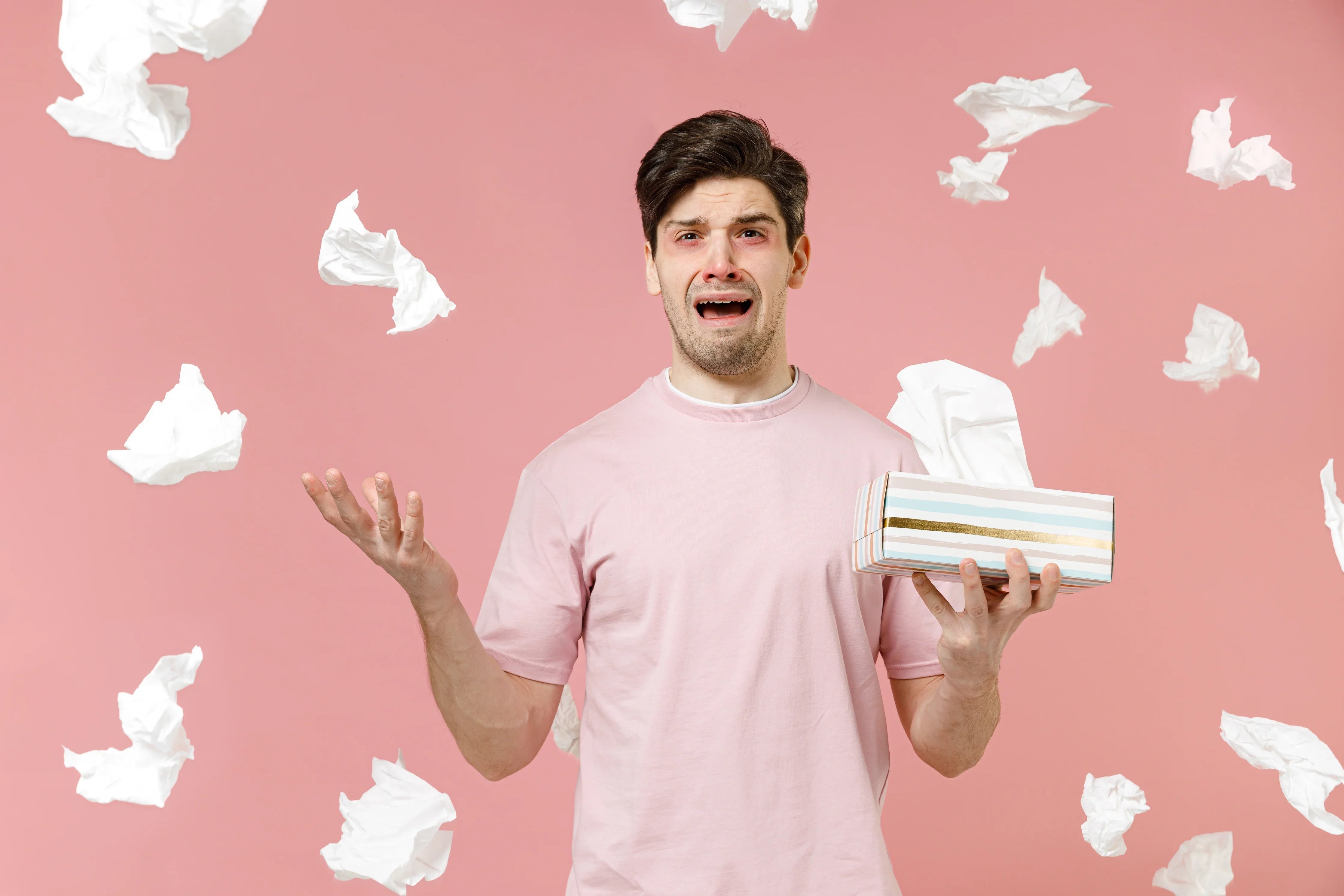 Man in pink shirt with red eyes holding tissues, surrounded by flying tissues, allergy concept