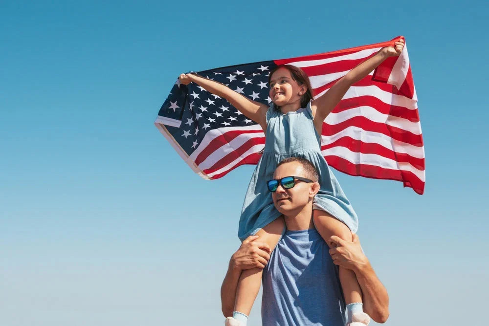 Father carrying daughter on shoulders, holding US flag, both in cotton clothing, blue sky background