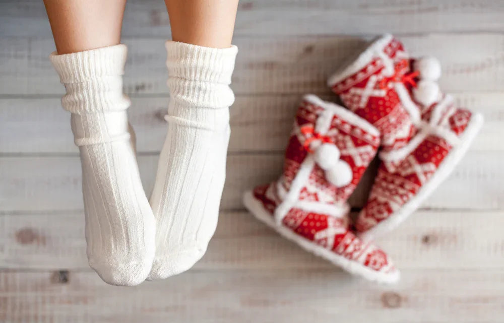 Pair of feet in white organic cotton socks beside red winter slippers on wooden floor