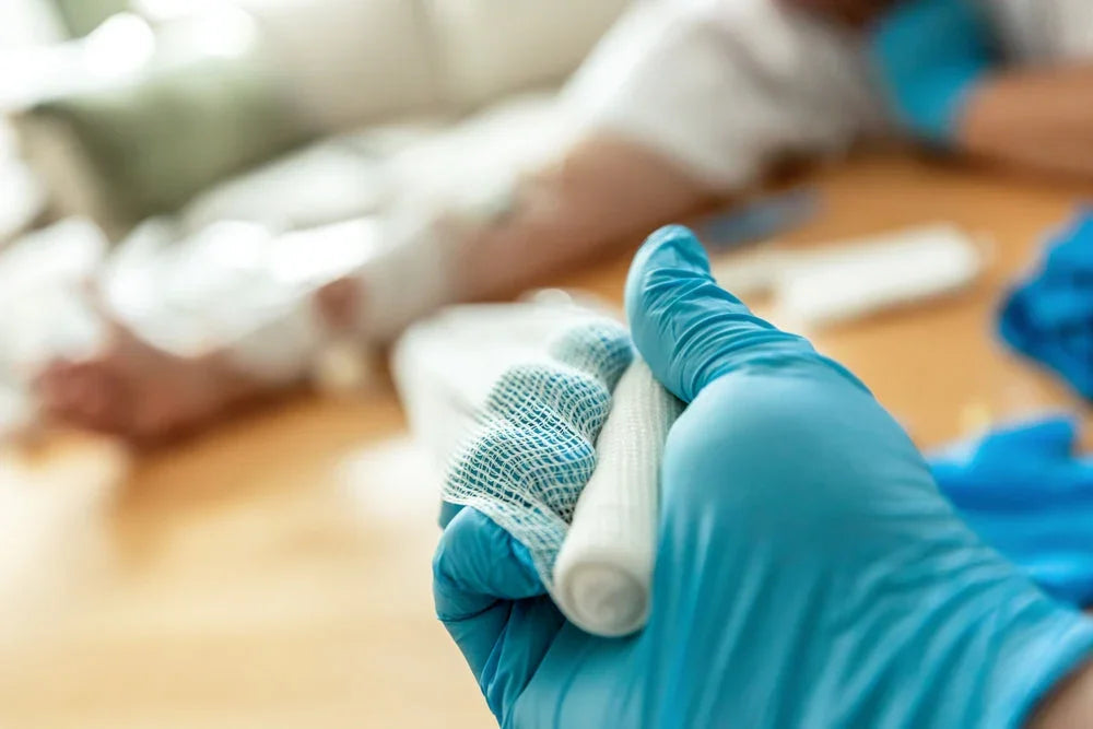 Gloved hand holding gauze bandage for sensitive skin, person in background on table