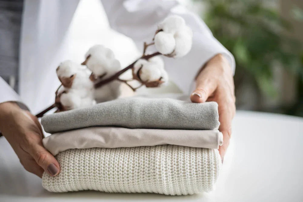 Hands holding folded organic cotton clothing and cotton branch, allergy-friendly fabrics