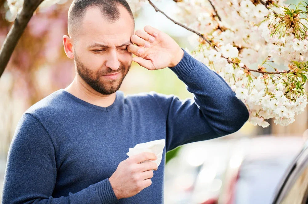 Man with tissue rubbing eye outdoors near blooming tree, allergy symptoms in spring