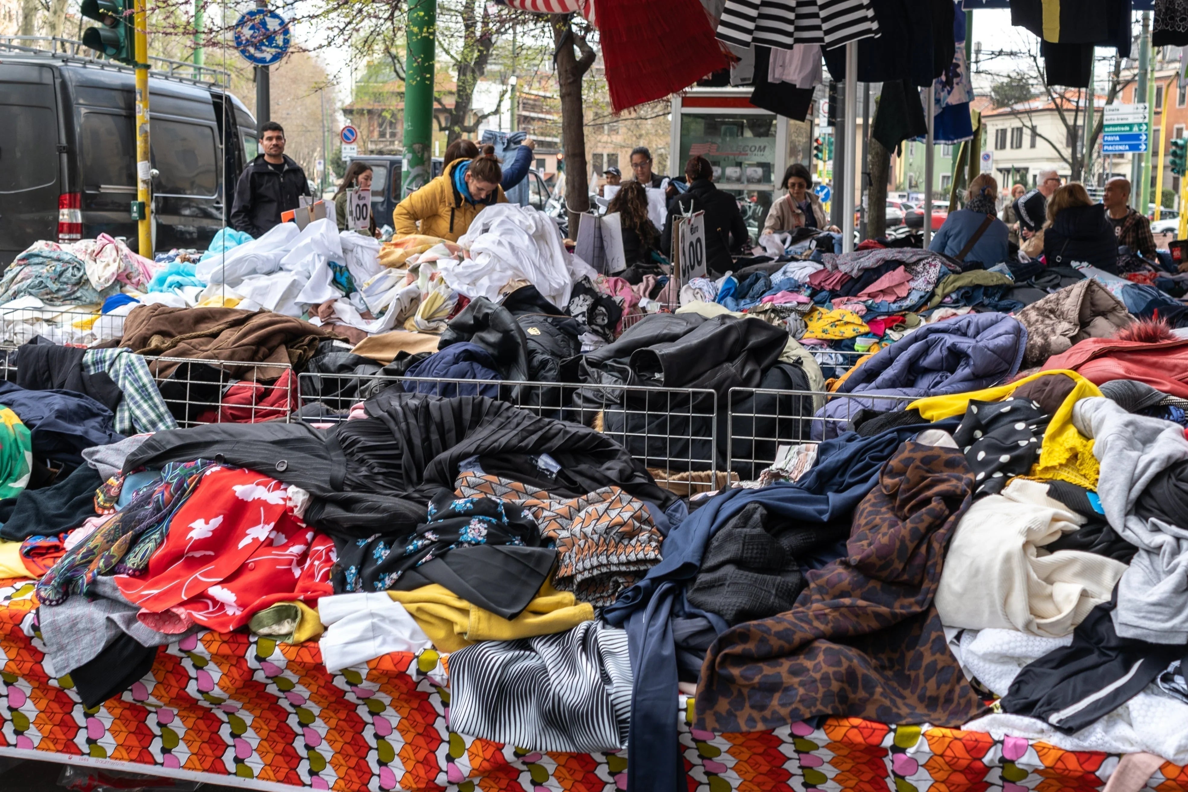 Outdoor market with assorted clothes piled on tables, shoppers browsing allergy-free apparel