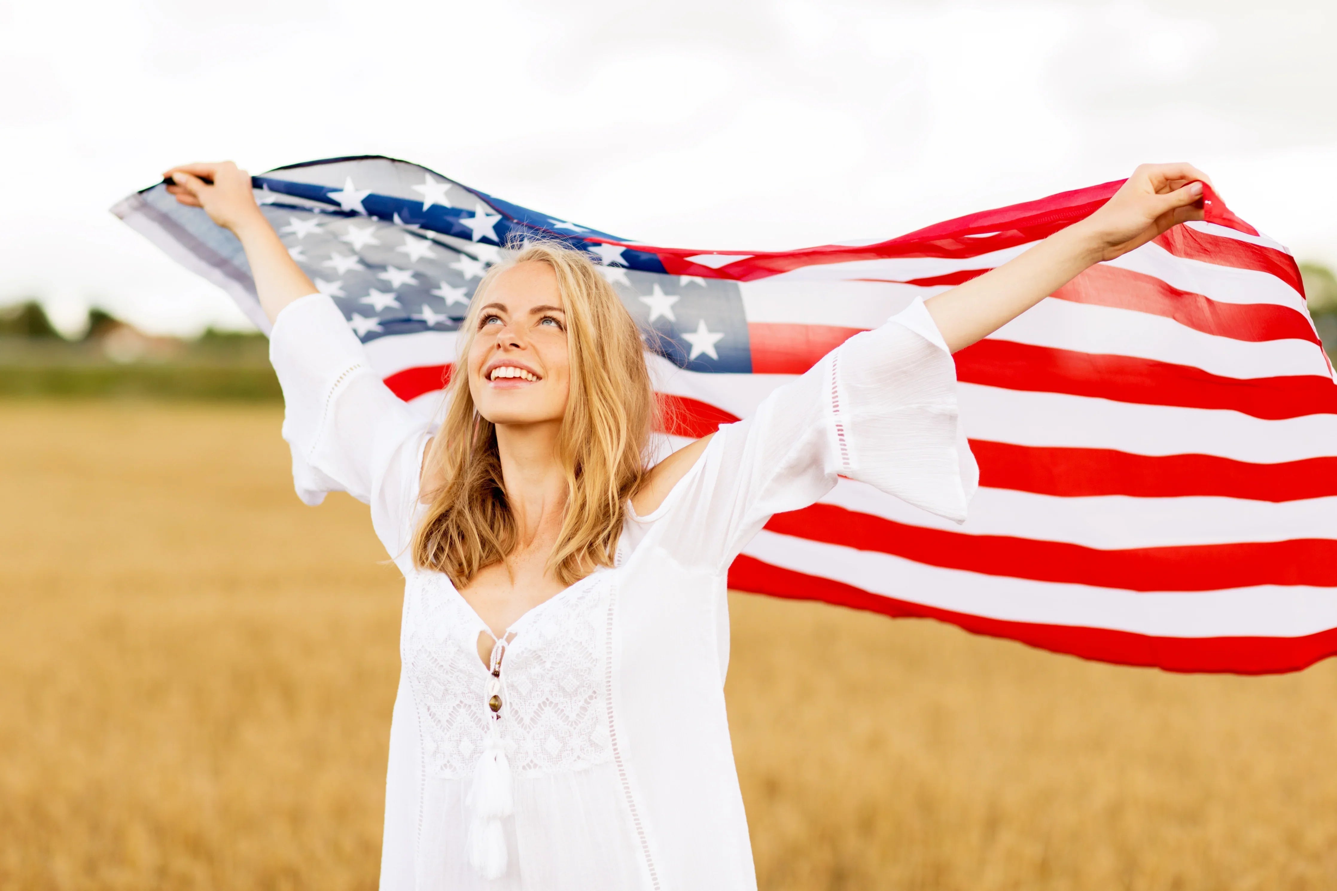 Smiling woman in a white cotton dress holding American flag in a field, allergy-free apparel
