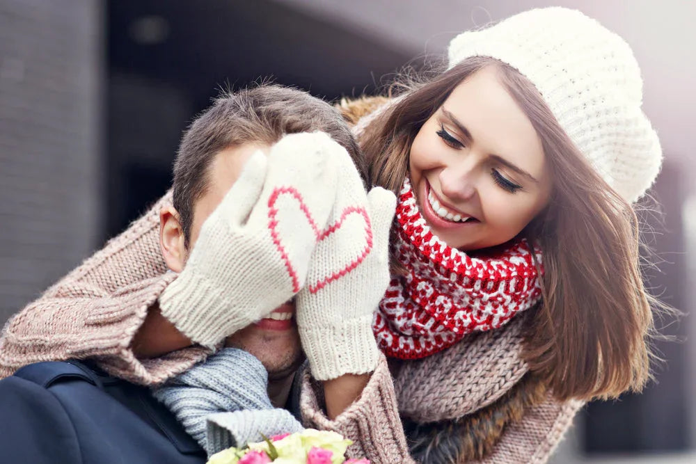 Smiling woman in organic cotton winter knitwear covers man's eyes with heart mittens outdoors