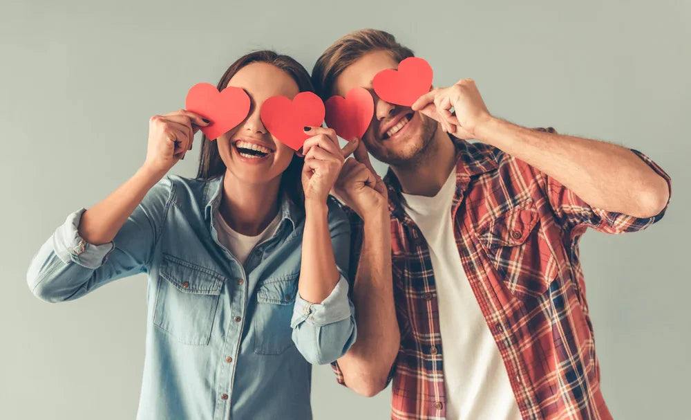 Smiling couple in cotton shirts holding heart cutouts over eyes, allergy-free apparel theme