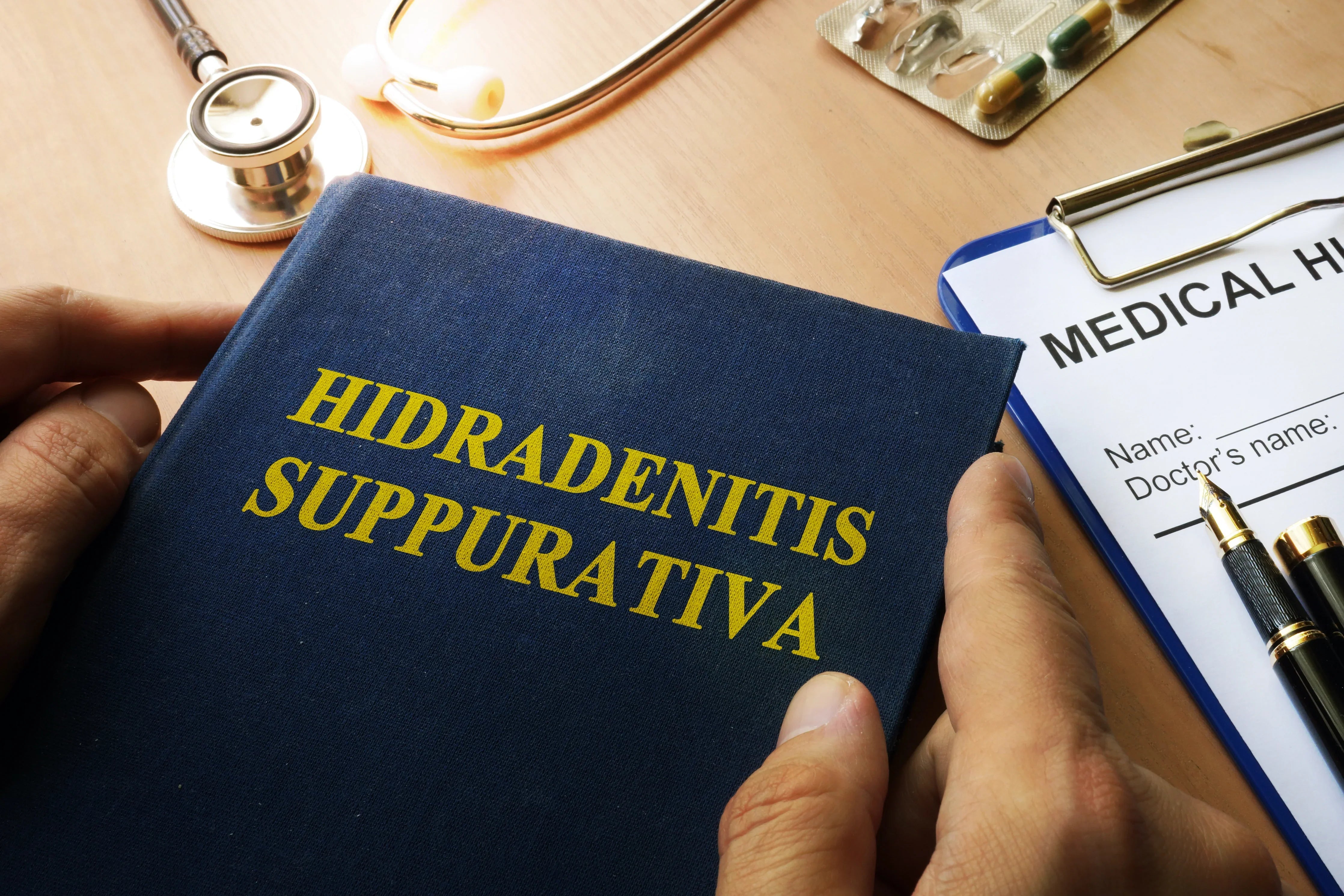 Book labeled Hidradenitis Suppurativa, stethoscope, medical form and pills on table, allergy-free apparel context