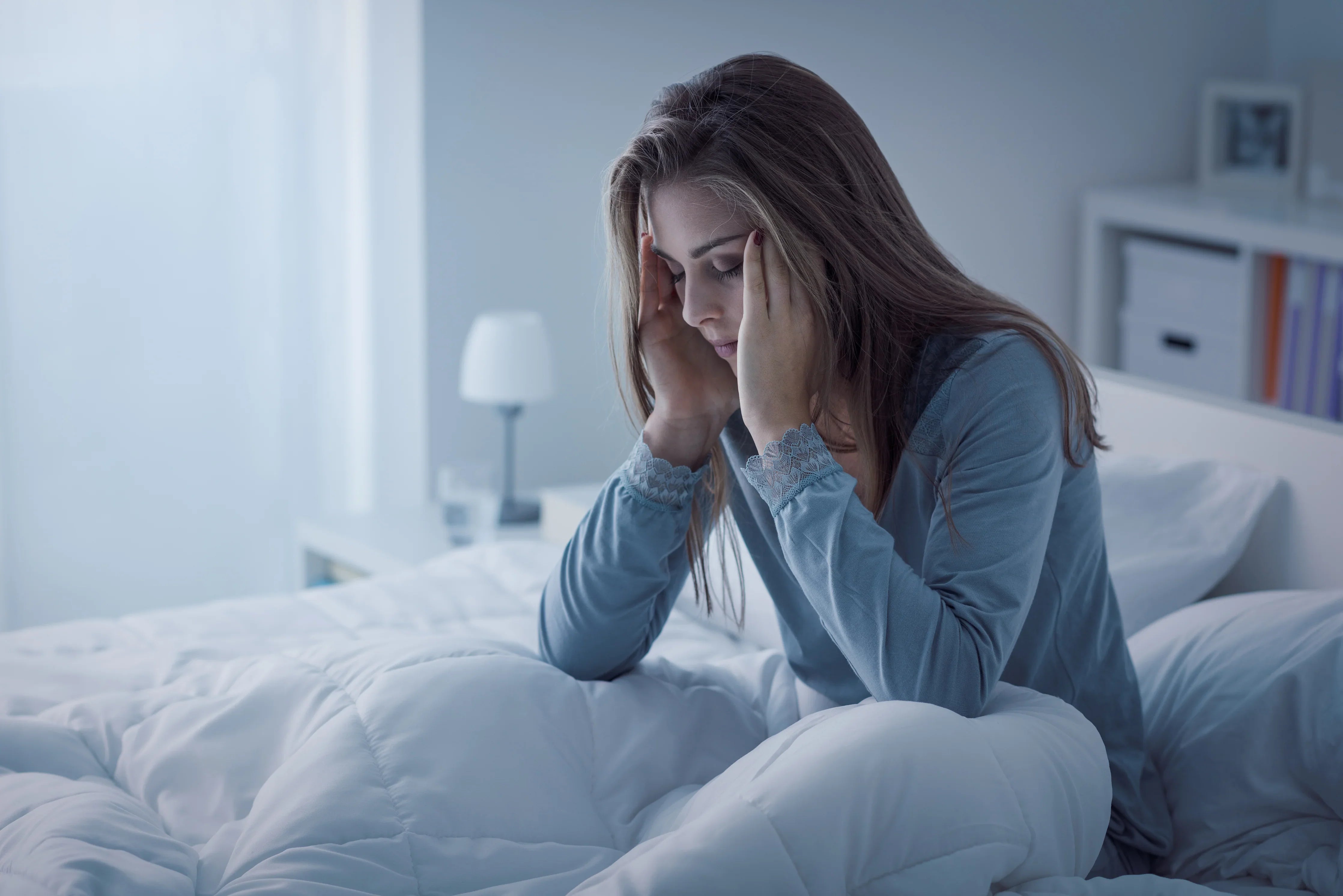 Woman in blue hypoallergenic pajamas sitting on bed, looking stressed in bright bedroom