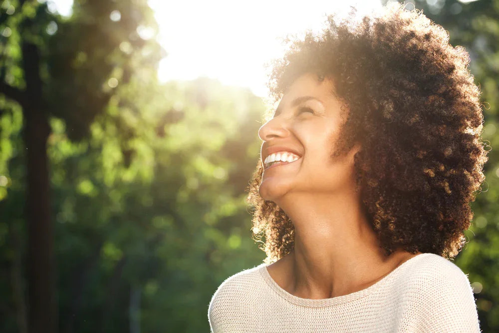 Smiling woman in light cotton sweater outdoors, sunlight and green trees in background