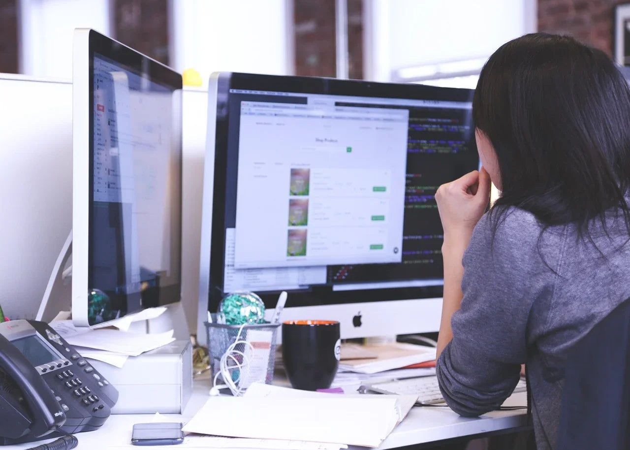 Person working at a desk with dual monitors and office supplies, viewing product listings online.